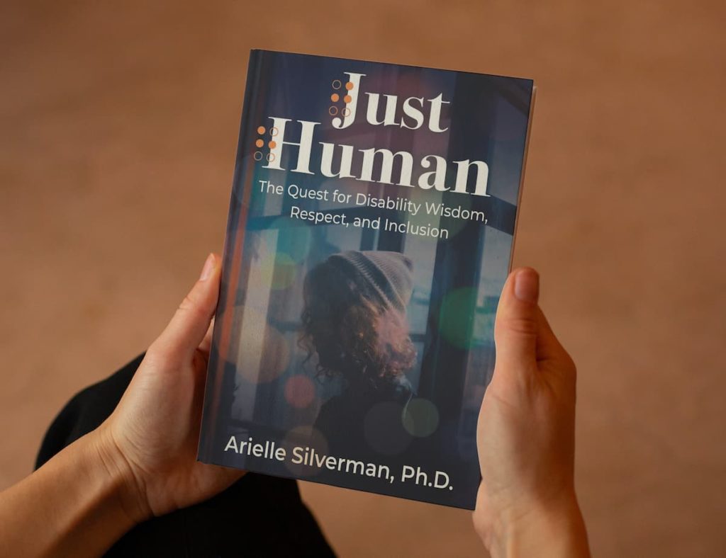 Two hands holding a physical copy of the book "Just Human: The Quest for Disability Wisdom, Respect, and Inclusion" by Arielle Silverman, Ph.D. The cover is of a young adult with brown curly hair and a beanie, standing with their back to a window with colorful circular light reflections surrounding them. Below the picture, it gives the author name, Arielle Silverman, Ph.D.